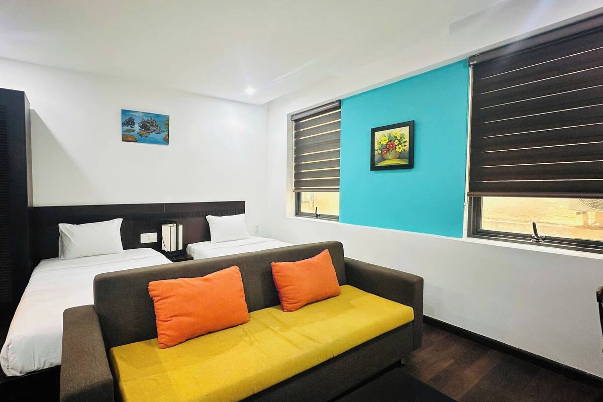 A hotel room with a bed, a sofa, a window with blinds, and a painting on the wall.