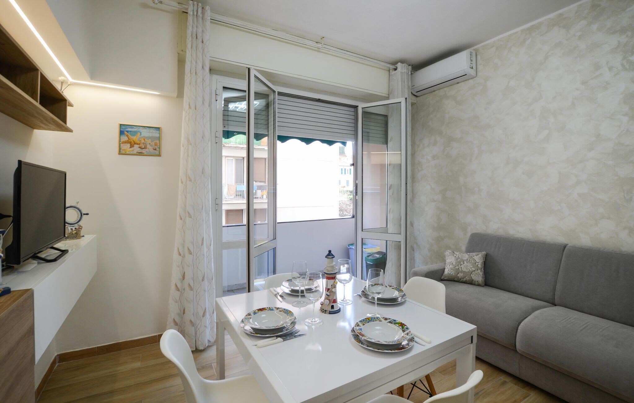Ein kleiner, gut beleuchteter Raum mit einem für vier Personen gedeckten Esstisch, einem grauen Sofa, einem Fernseher und einem Fenster mit Blick auf Gebäude.