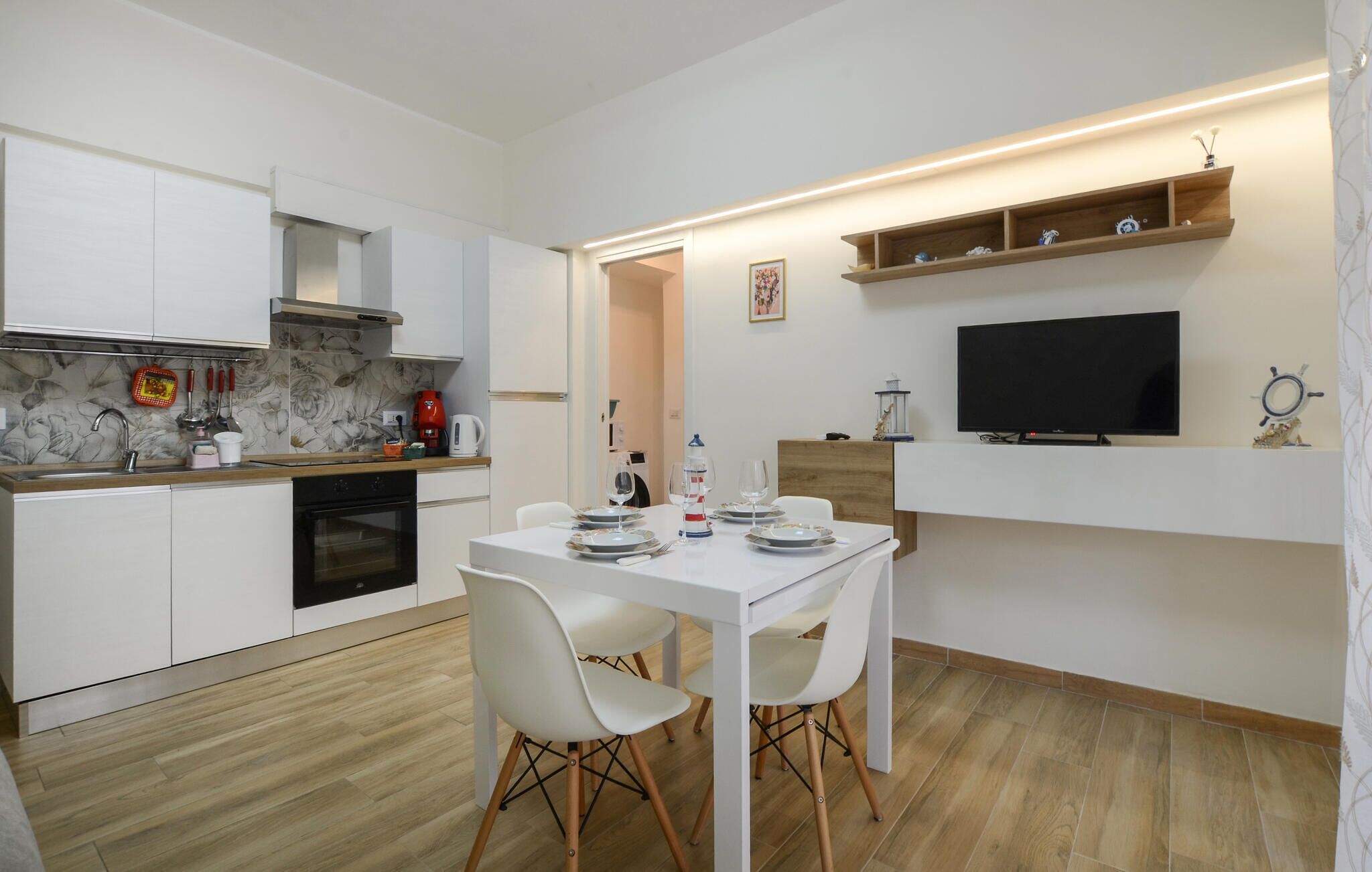Una cucina compatta con mobili bianchi, forno integrato e zona pranzo con tavolo e sedie bianche.
