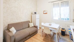 Een gezellig, compact leefruimte met een bank, een kleine tafel met stoelen, een koelkast en een raam met zonwering.