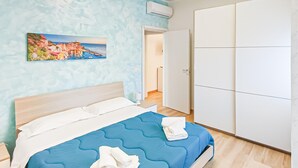 Een slaapkamer met een bed, een schilderij van een kustlandschap, een kledingkast en een deur naar een andere kamer.