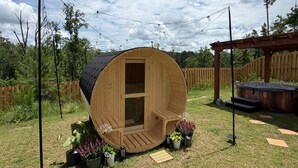 Een houten, vatvormige constructie met een deur, omringd door potplanten, een pergola en een houten terras.