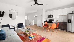 Ein modernes Wohnzimmer mit Sofa, Couchtisch und Sesseln, ein Essbereich mit Tisch und Stühlen, eine Küche mit Schränken und Geräten sowie ein Deckenventilator.