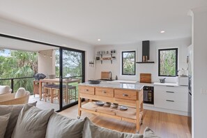 A modern kitchen with a large island, wooden cabinets, and a dining area with a glass door leading to a balcony. - The Reid Residence - Merimbula (Merimbula)