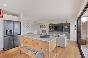 A modern kitchen with a built-in refrigerator, a wooden dining table with plates, a grey sofa, and a TV in the living area. - The Reid Residence - Merimbula (Merimbula)
