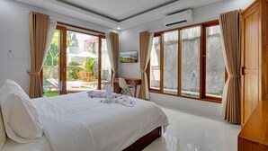 A hotel room with a large bed, two windows with curtains, a small table with chairs, and an air conditioning unit. - Sedana Villa Sanur (Denpasar)