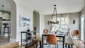 Un comedor con una mesa de madera, sillas y una araña. Se ve una escalera que sube a otro nivel, y una zona de cocina al fondo.