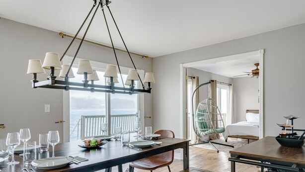 Comedor con mesa de madera preparada para cuatro personas, lámpara colgante y vista al océano a través de grandes ventanales.