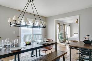 Uma sala de jantar com uma mesa de madeira posta para quatro pessoas, um lustre pendurado e vista para o oceano através de grandes janelas.