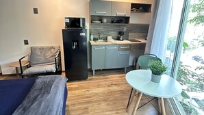 Ett kompakt hotellrum med en säng, ett litet pentry, en matplats med bord och stolar samt ett fönster med utsikt över omgivningen.