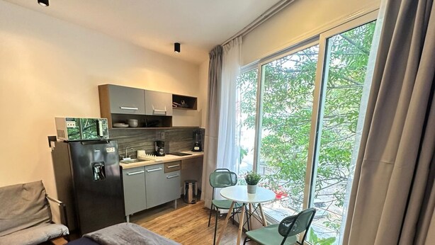 Ett modernt hotellrum med en säng, ett litet pentry, ett matbord med stolar och ett stort fönster med gardiner.