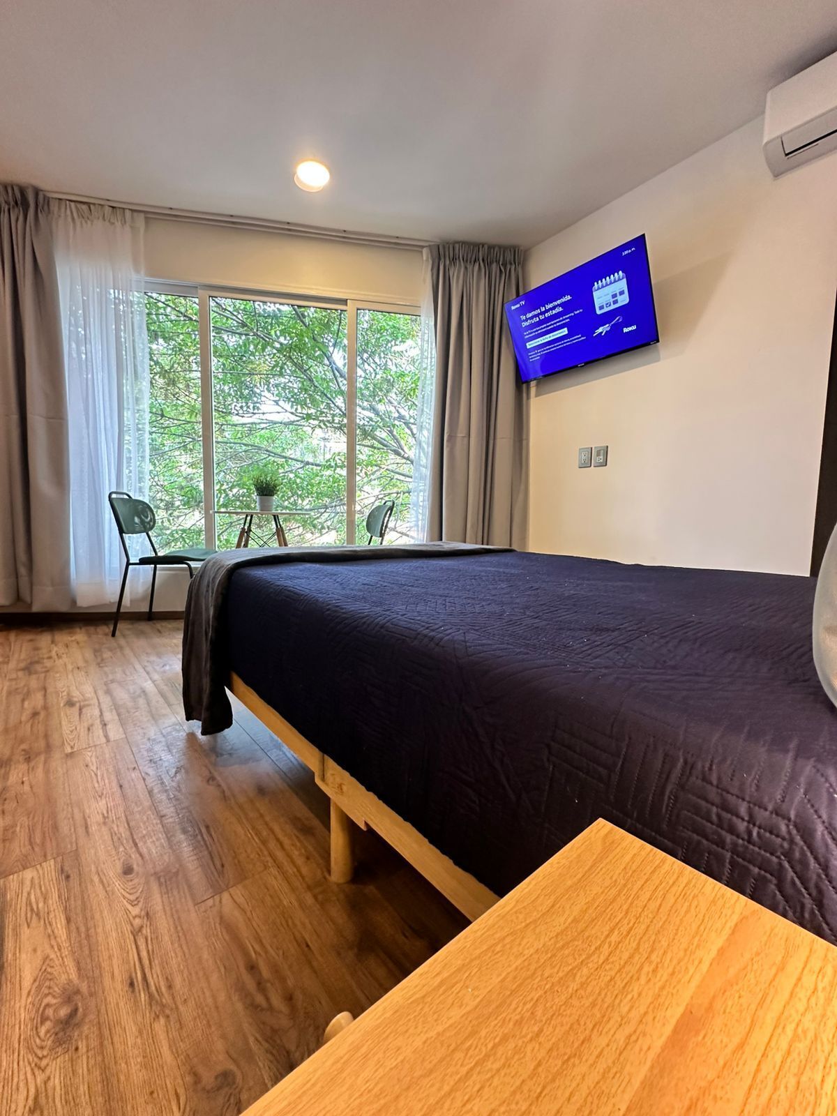 A hotel room with a bed, a TV mounted on the wall, a window with curtains, and a small table with chairs.