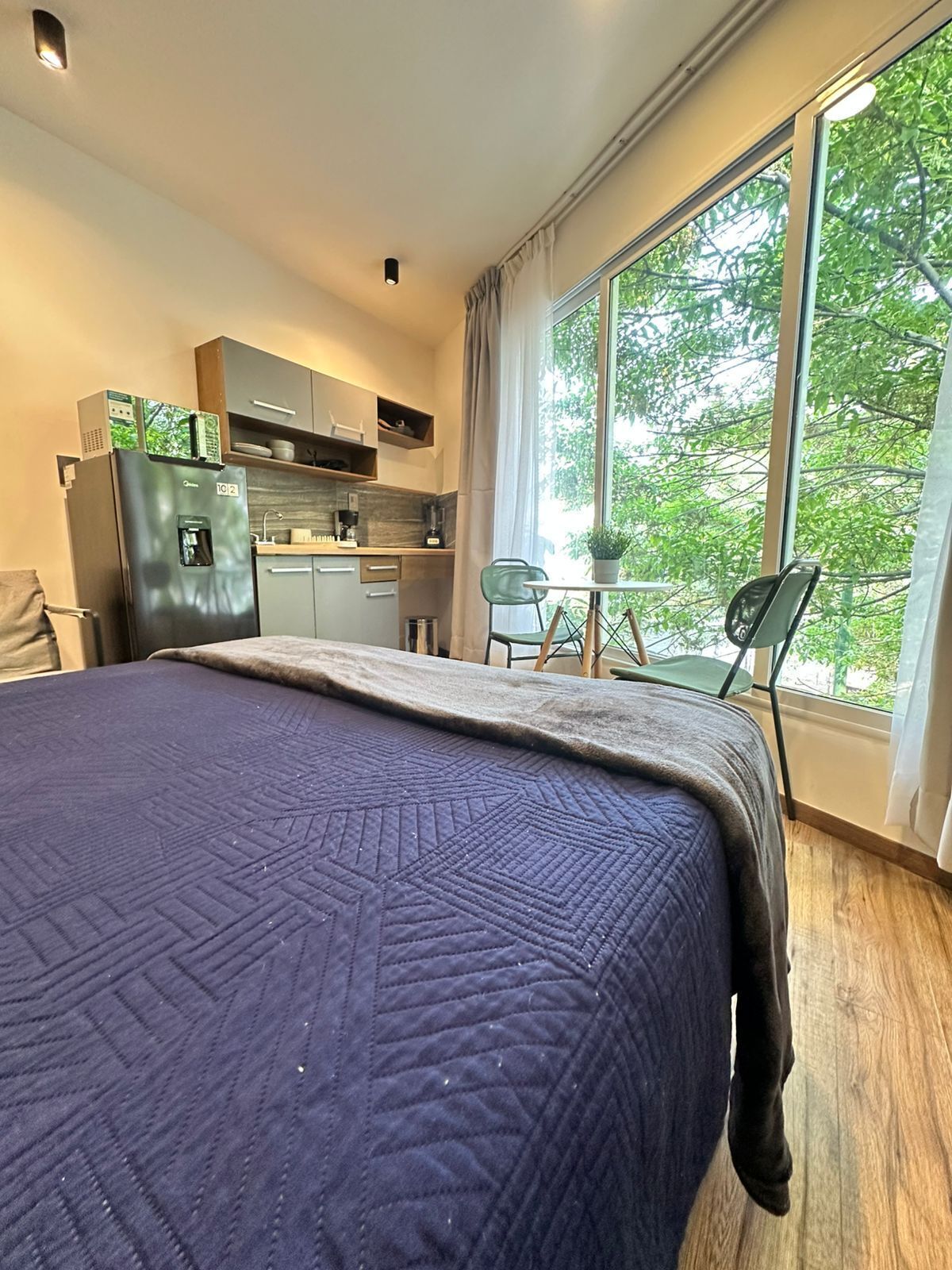 A compact room with a bed, kitchenette, and a window with a view of trees.