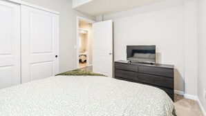 Une chambre à coucher avec un lit, une commode et un téléviseur. On peut voir des portes blanches et un couloir.