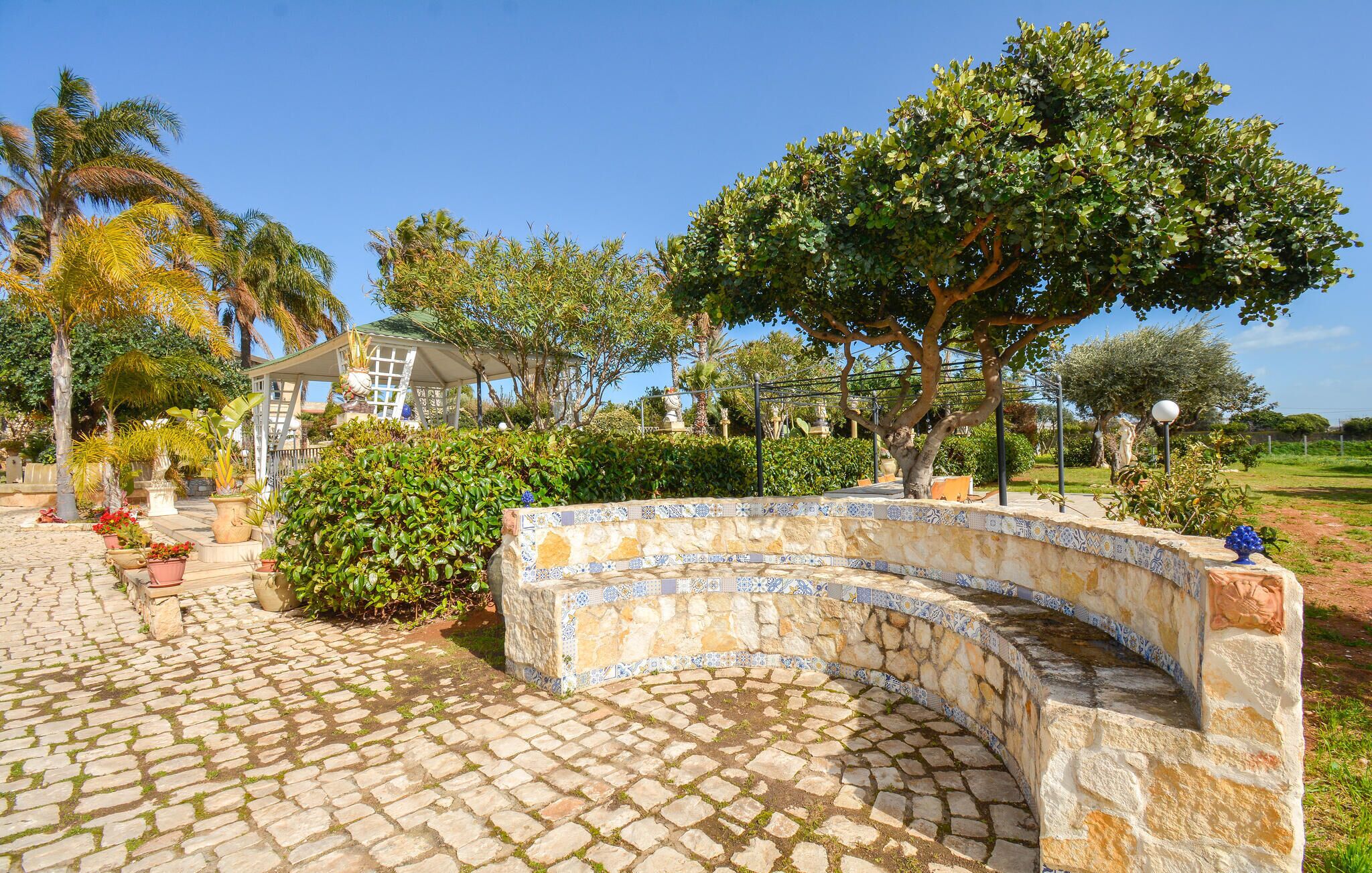 A stone pathway leading to a curved stone bench with a tiled border, surrounded by lush greenery and a clear blue sky.