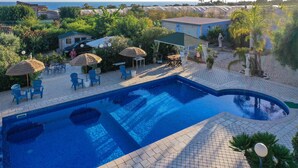 Una piscina con azulejos azules, rodeada de sillones de descanso y sombrillas, con vista al mar y edificios al fondo.