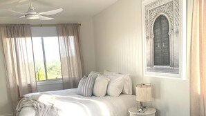 Ein Schlafzimmer mit einem großen Bett, einem kleineren Bett, einem Nachttisch mit Lampe, einem Deckenventilator und einem Fenster mit Vorhängen.