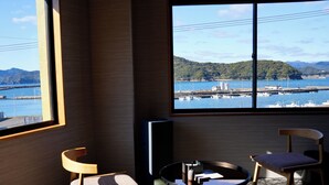 A room with a large window offering a view of a harbor, two wooden chairs, a small round table, and a flat-screen TV. - 1 and Only Hotel Io (Minamiise)