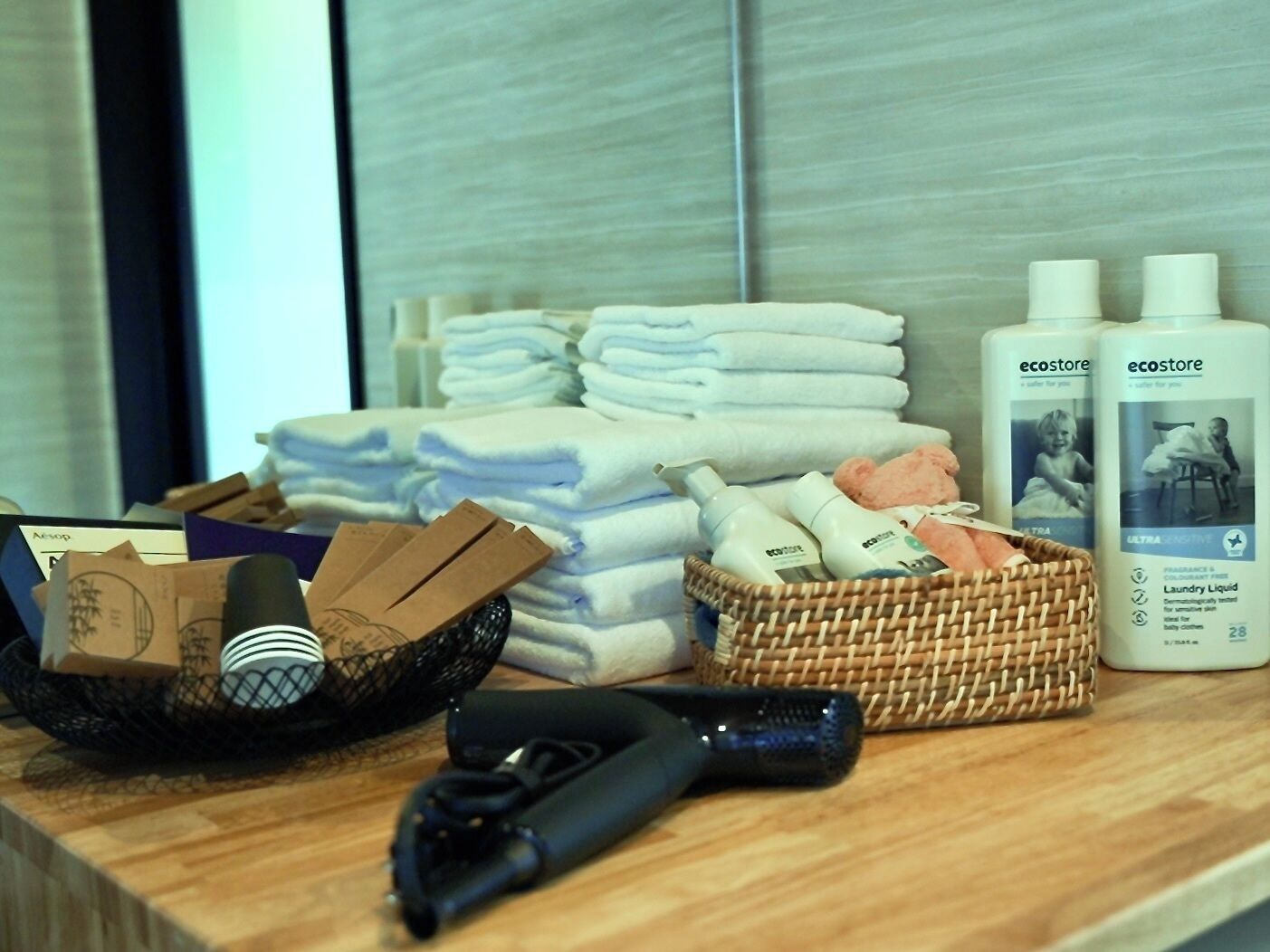 A hotel room with neatly stacked towels, bottles of Ecostore laundry liquid, a basket with toiletries, and a hairdryer on the counter.