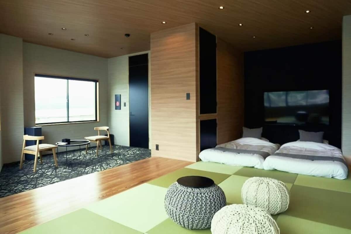 A modern hotel room with a large bed, a seating area with chairs, a small table, and a large window.