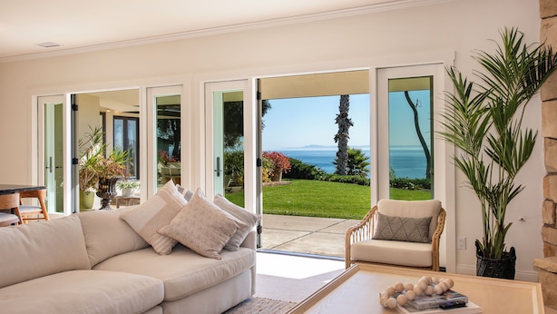 A modern living room with a beige sofa, a wooden coffee table, and a view of a garden and ocean through large glass doors. - Beautiful Home above Mesa Bluffs (Santa Barbara)