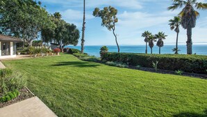 A well-maintained lawn with a view of the ocean, palm trees, and a house with a covered patio. - Beautiful Home above Mesa Bluffs (Santa Barbara)