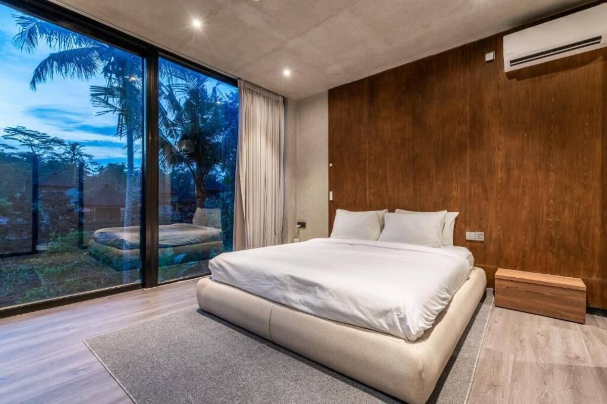 A modern bedroom with a large bed, a wooden accent wall, and a sliding glass door leading to a balcony with a view of palm trees.