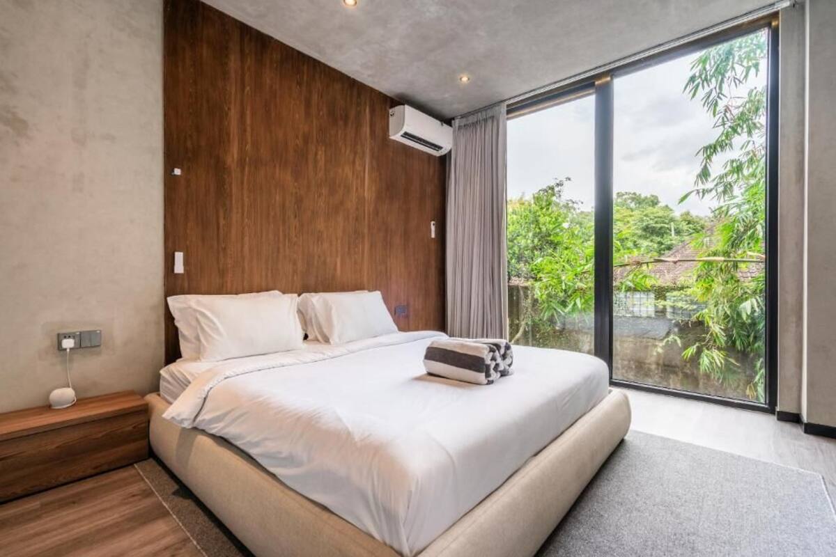 A modern bedroom with a large bed, wooden headboard, and a view of greenery through a sliding glass door.