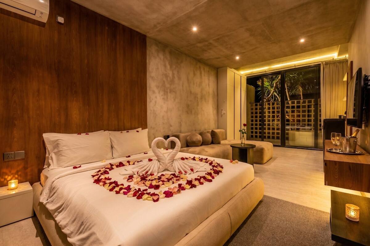 A modern bedroom with a large bed, a sofa, a TV, and a balcony with a view.