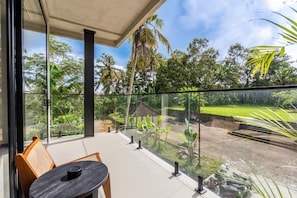 A balcony with a glass railing overlooking a garden with palm trees and a small structure.