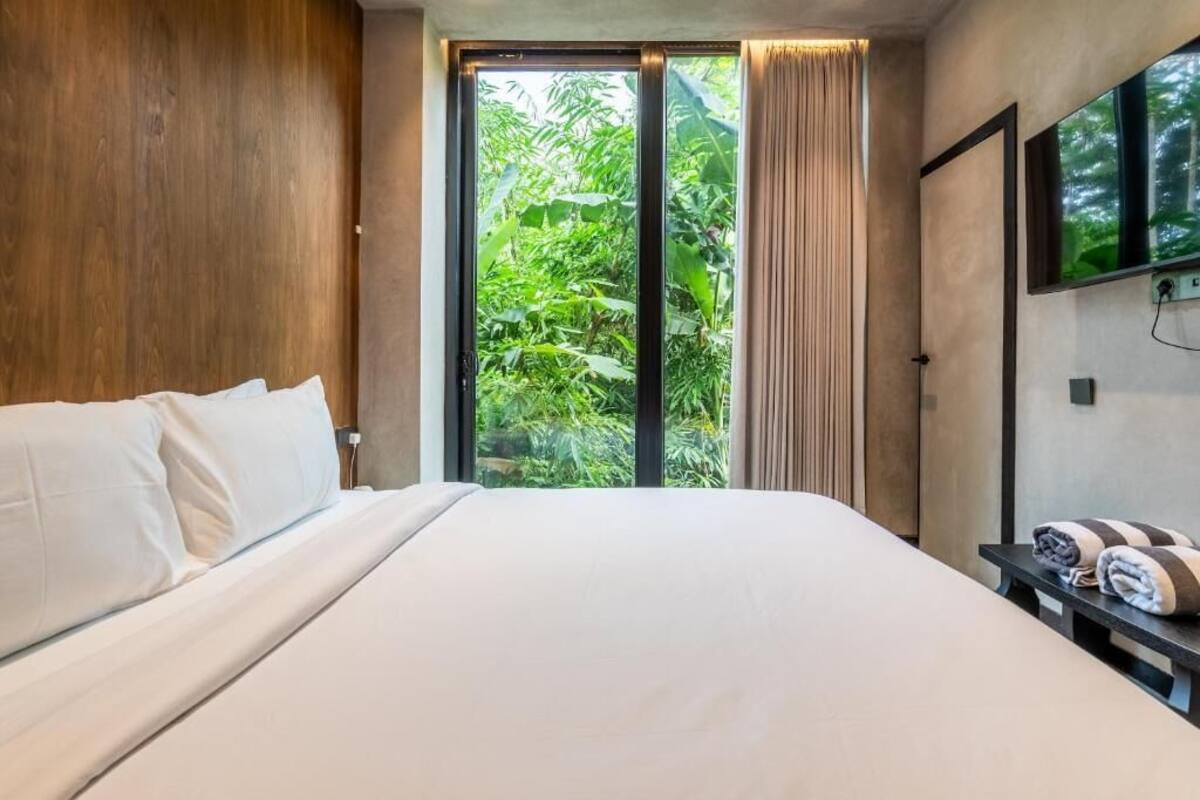A hotel room with a large bed, a TV mounted on the wall, and a sliding glass door to a lush garden.