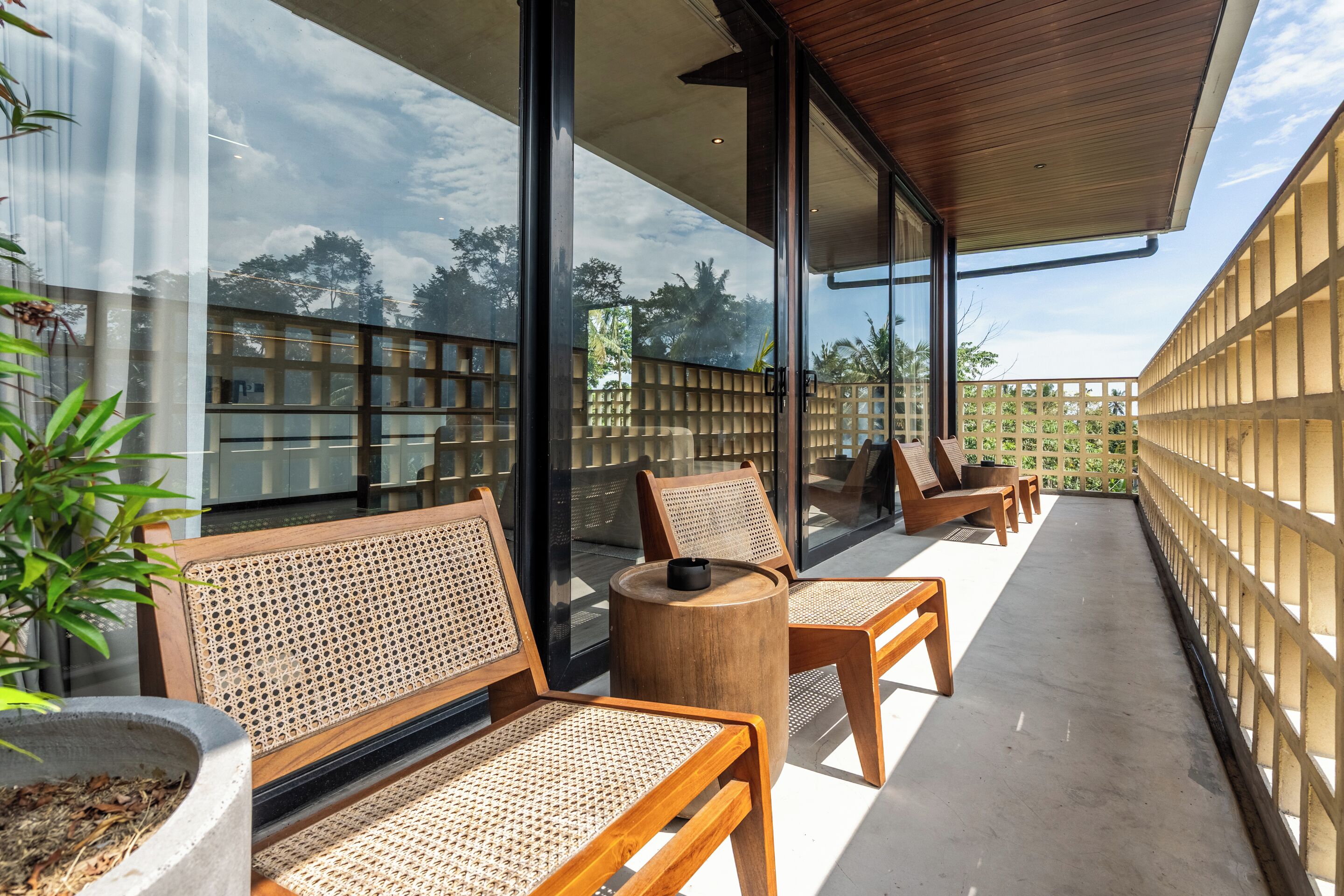 Un balcon avec des chaises en bois et une table ronde, de grandes fenêtres en verre et une vue sur les arbres.