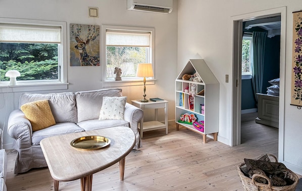 A cozy living room with a sofa, a side table, a lamp, a bookshelf, and a view of the outdoors.