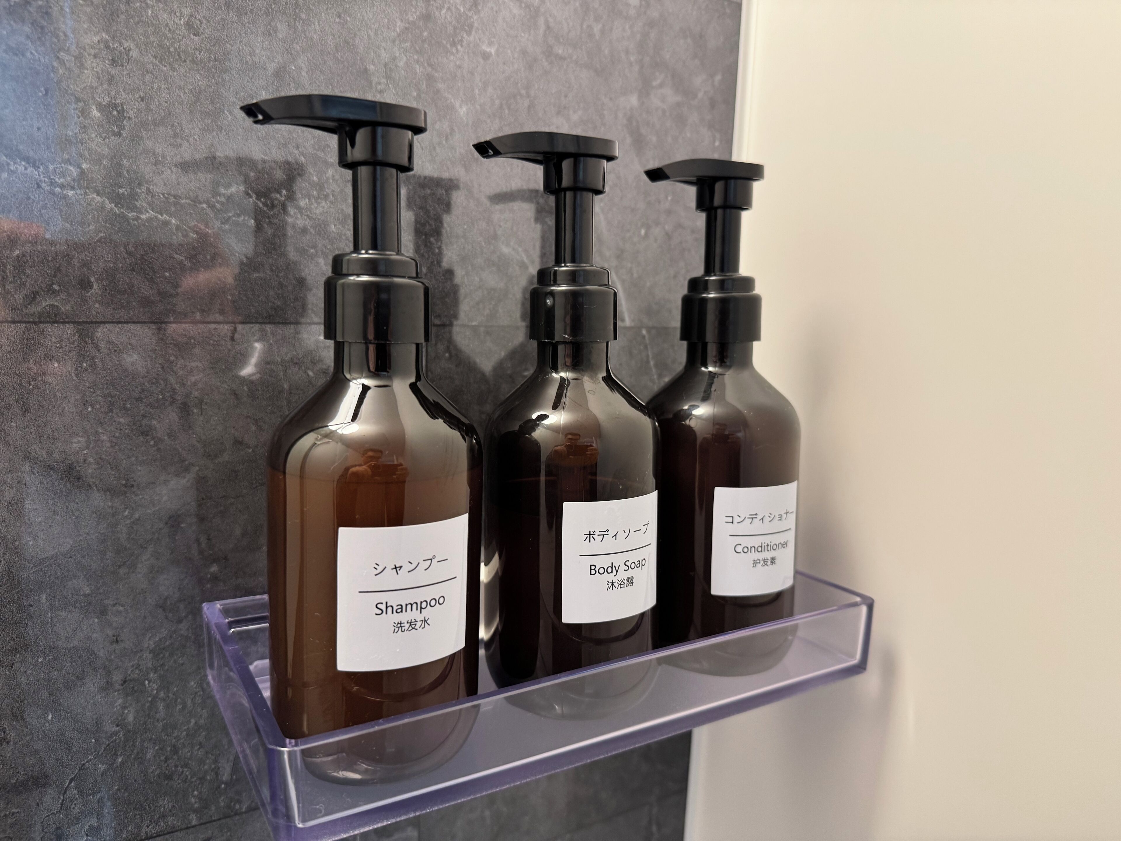 Three bottles of shampoo, body soap, and conditioner on a transparent shelf against a textured wall.
