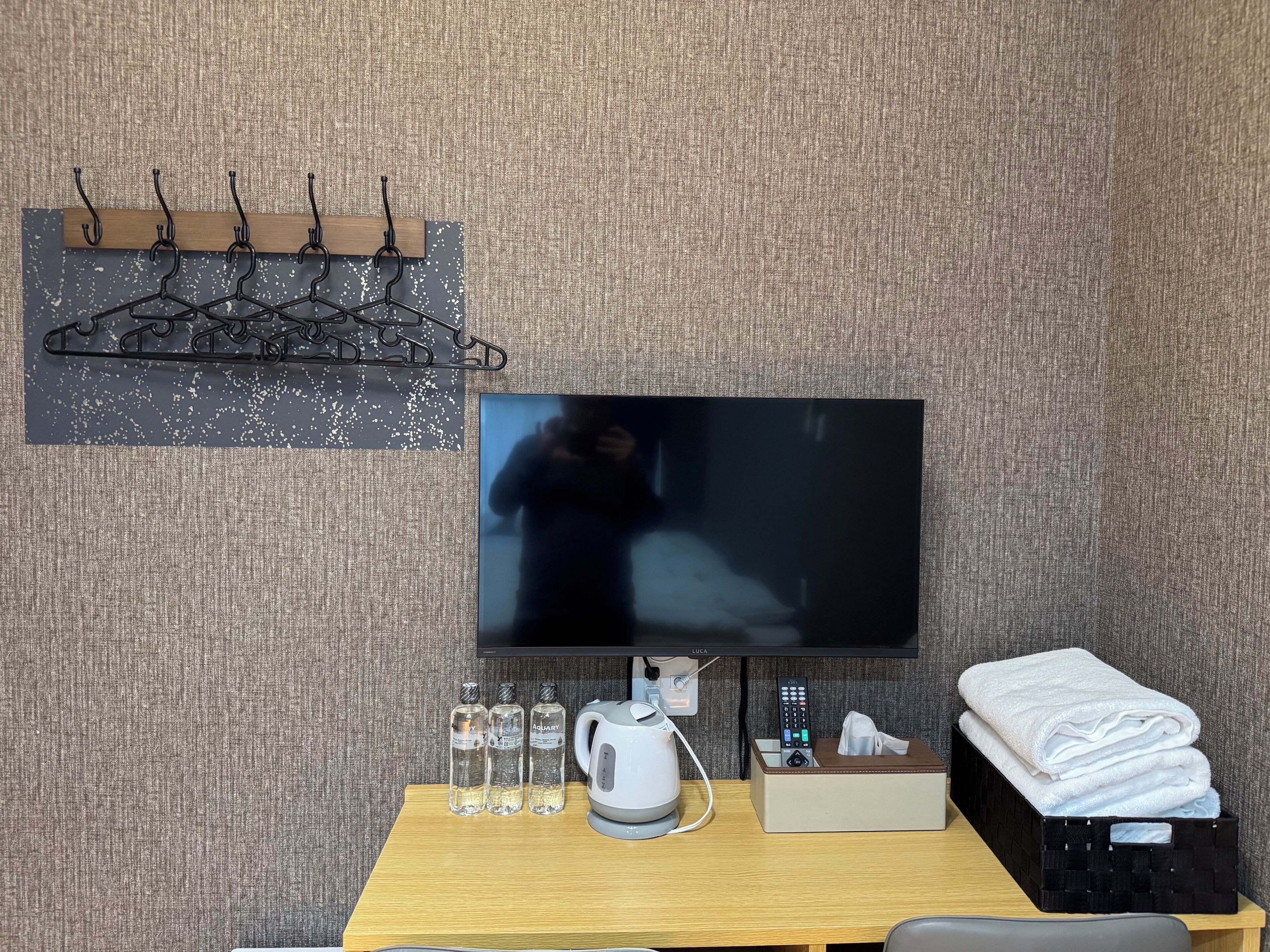 A hotel room with a flat-screen TV, a wooden table with a kettle, glasses, and a remote control.