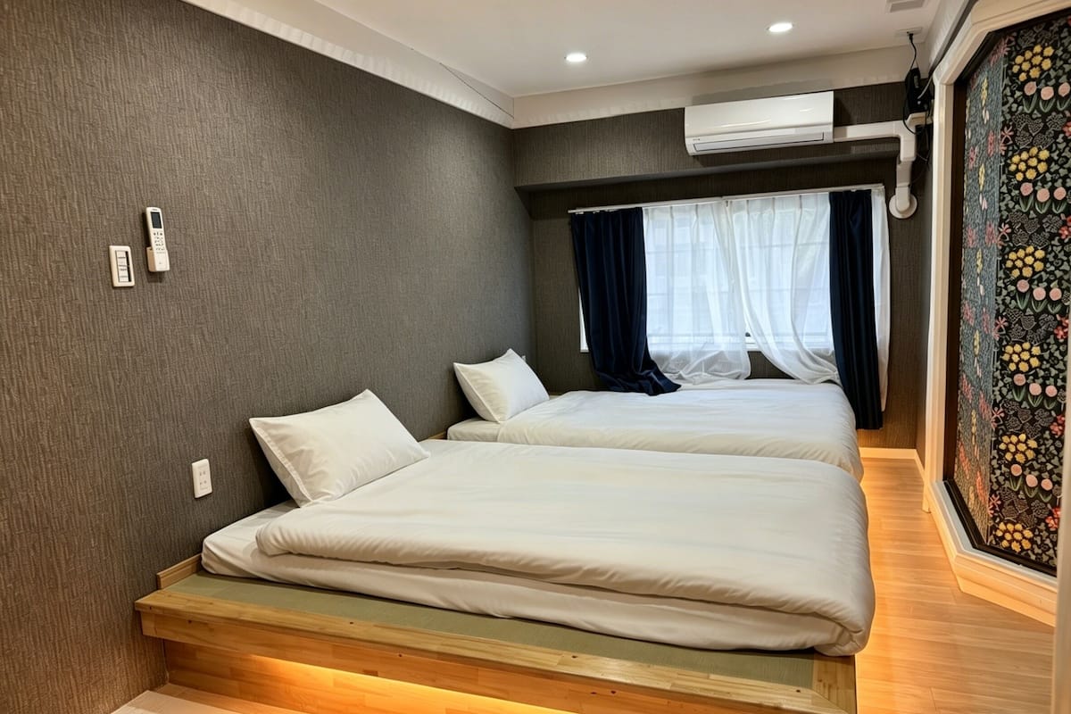 Habitación de hotel con dos camas, un banco de madera, una ventana con cortinas traslúcidas y un equipo de aire acondicionado.