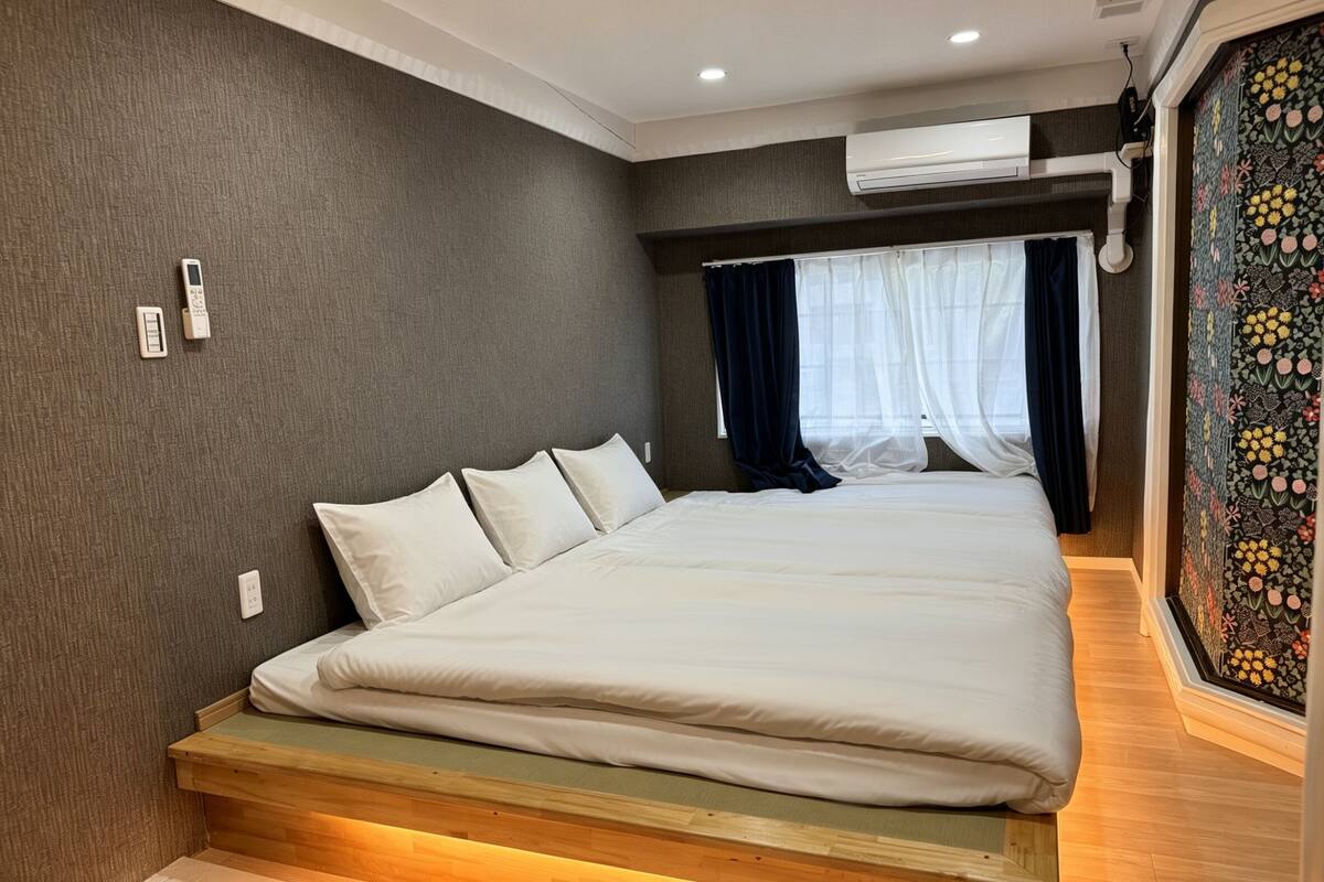 A bedroom with a large bed, a wooden bench at the foot of the bed, a wall-mounted air conditioner, and a window with sheer curtains.