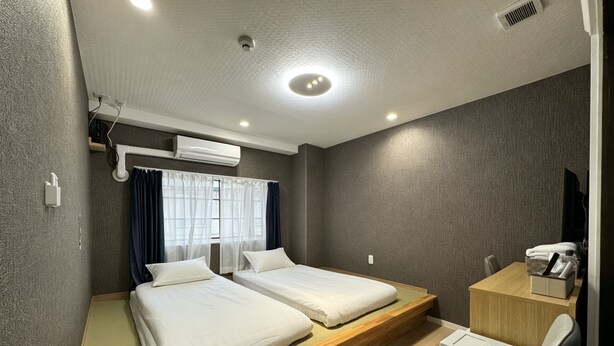 Quarto de hotel com duas camas, uma escrivaninha, uma cadeira e uma janela com cortinas.