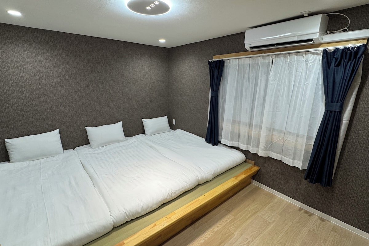 A bedroom with a bed, grey walls, a window with curtains, and an air conditioning unit.