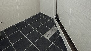 Um espaço de chuveiro pequeno e fechado, com paredes e chão revestidos de azulejos, um ralo e uma única luminária no teto.