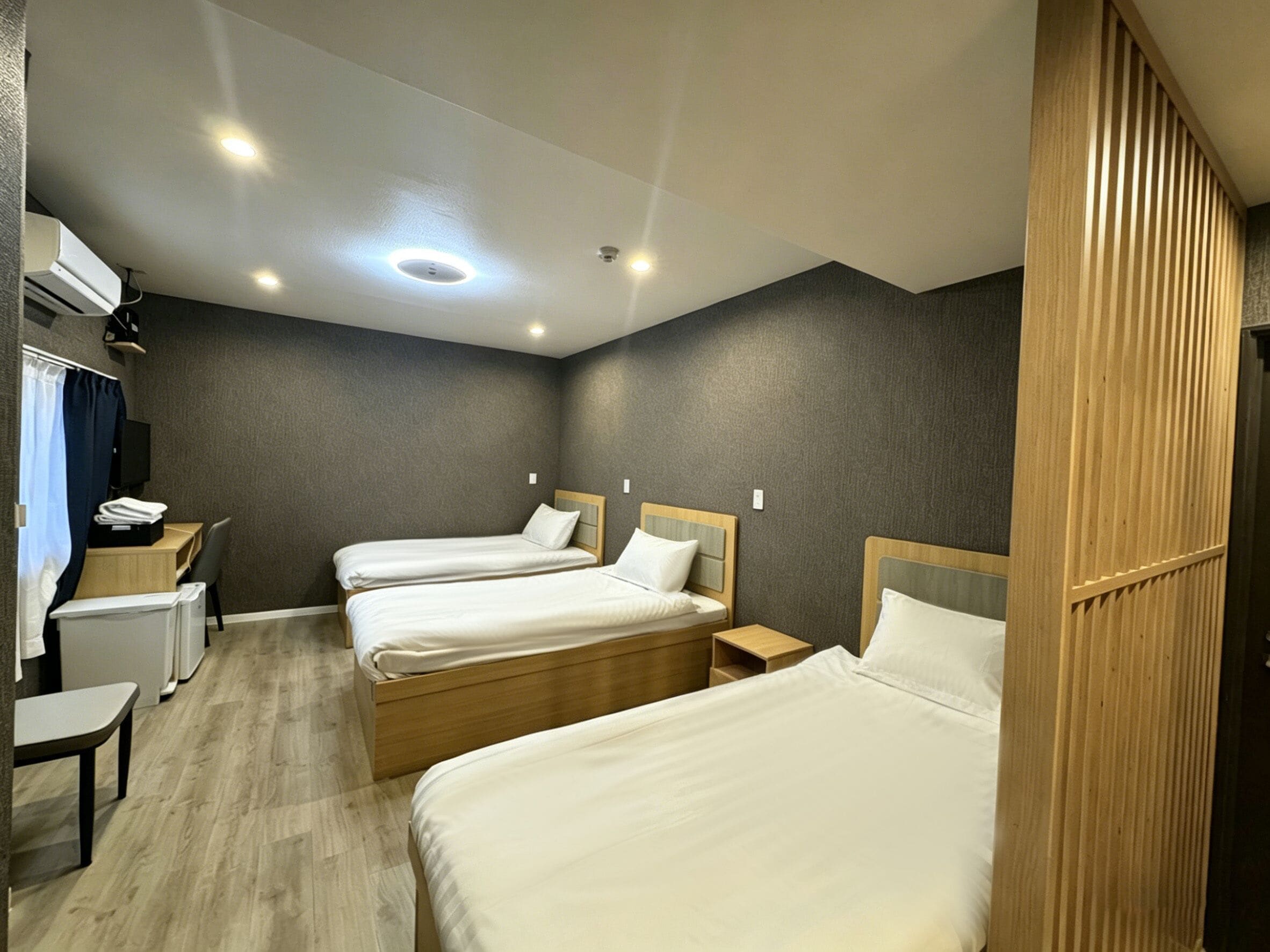 Quarto de hotel com três camas de solteiro, uma escrivaninha, uma cadeira e uma mesinha.