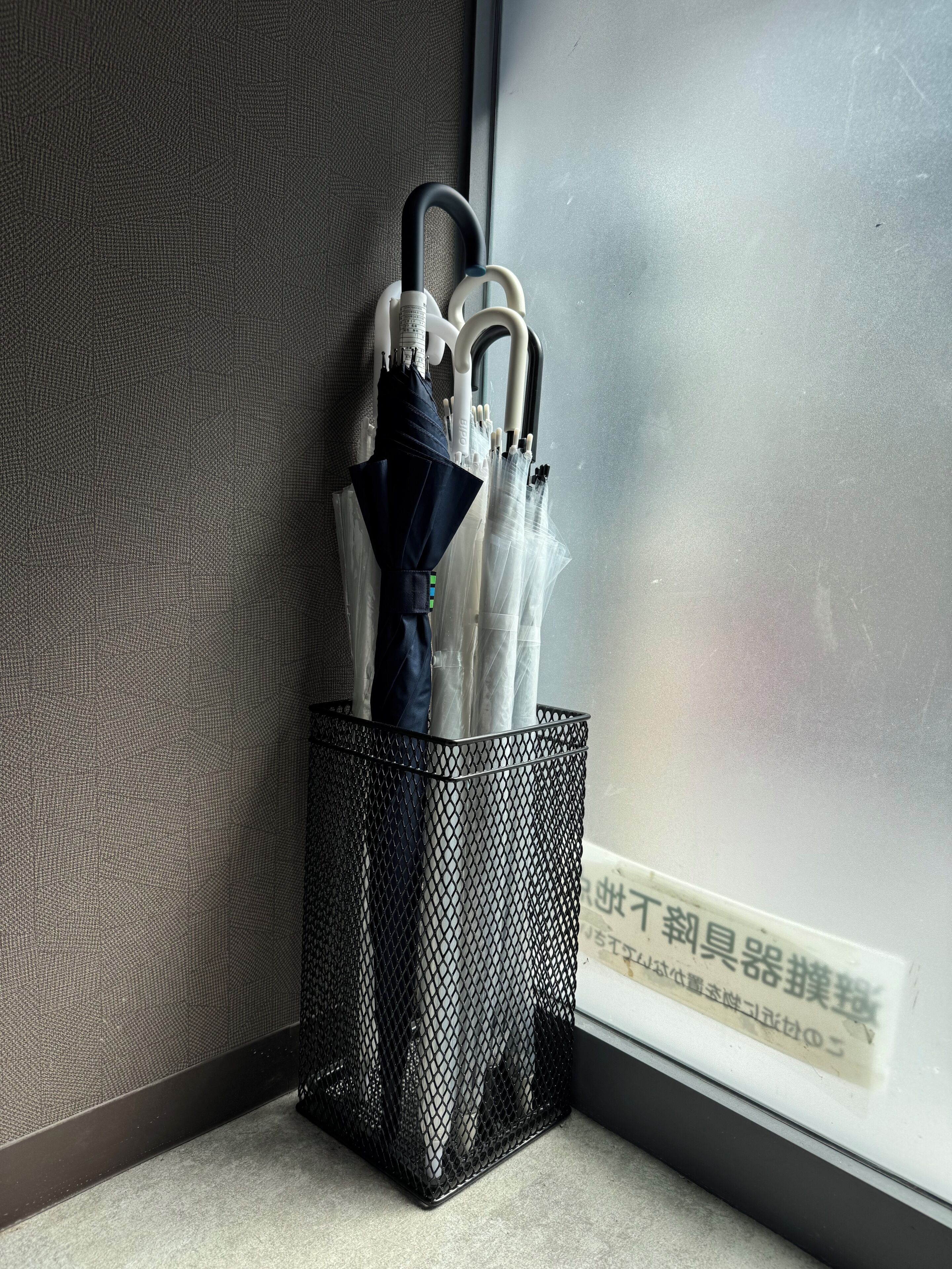 A metal mesh container holding umbrellas near a window with a textured wall.