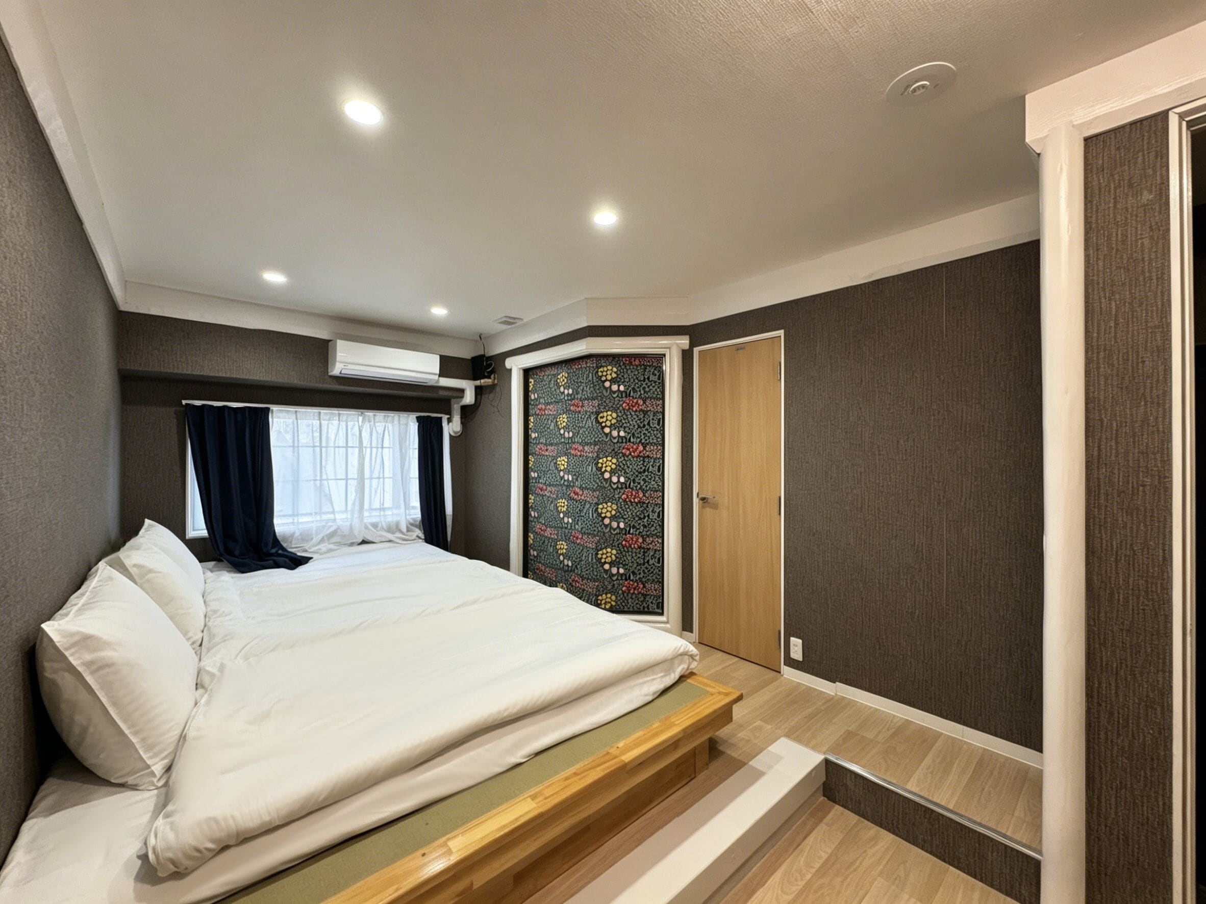 Un dormitorio con cama, ventana con cortinas, puerta con papel tapiz estampado y unidad de aire acondicionado integrada.