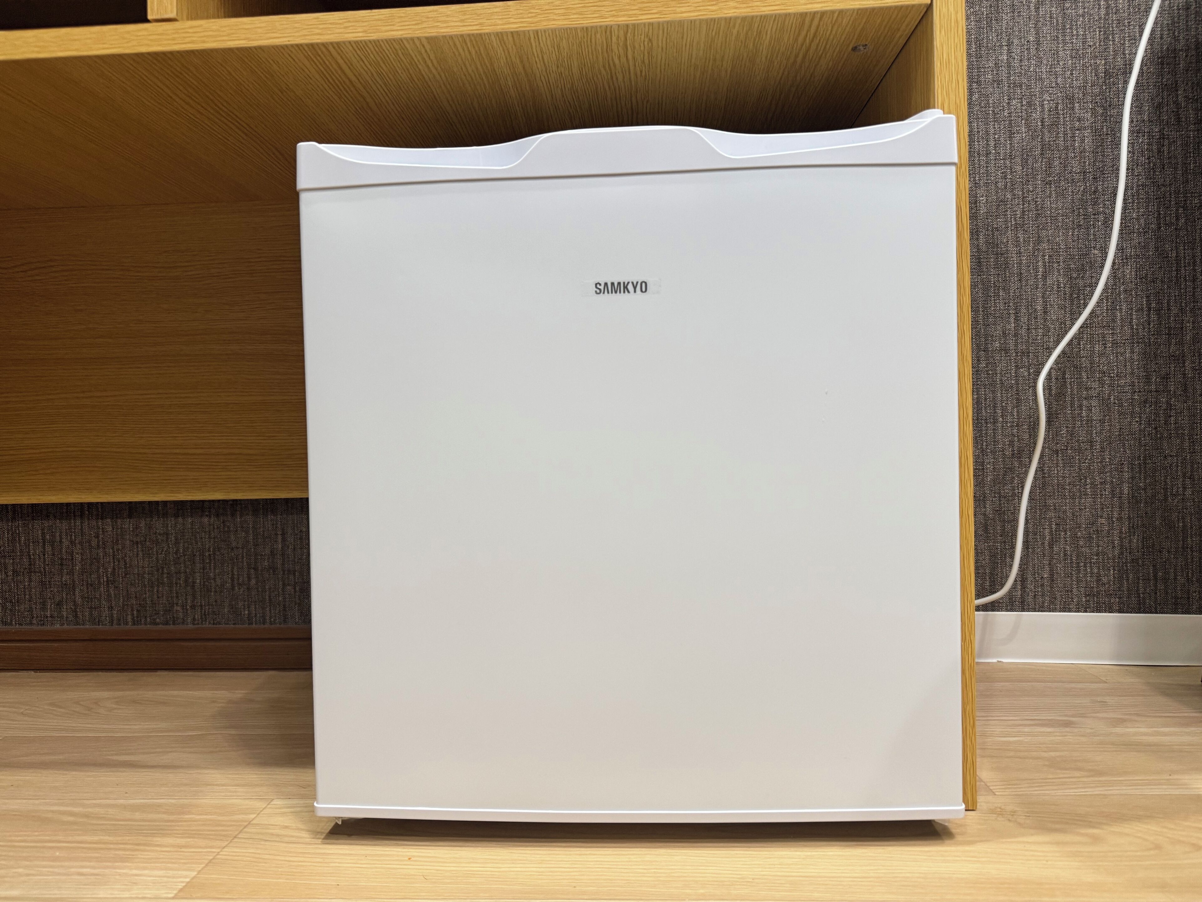 A white refrigerator with the brand name 'SAMKYO' on the front, placed on a wooden floor with a textured wall in the background.