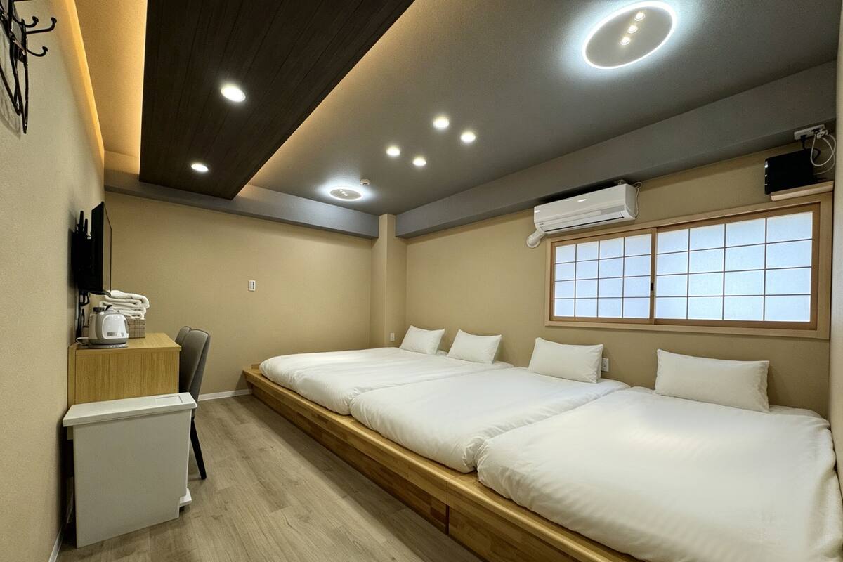 A hotel room with a large bed, a small desk with a kettle, a chair, and an air conditioning unit.