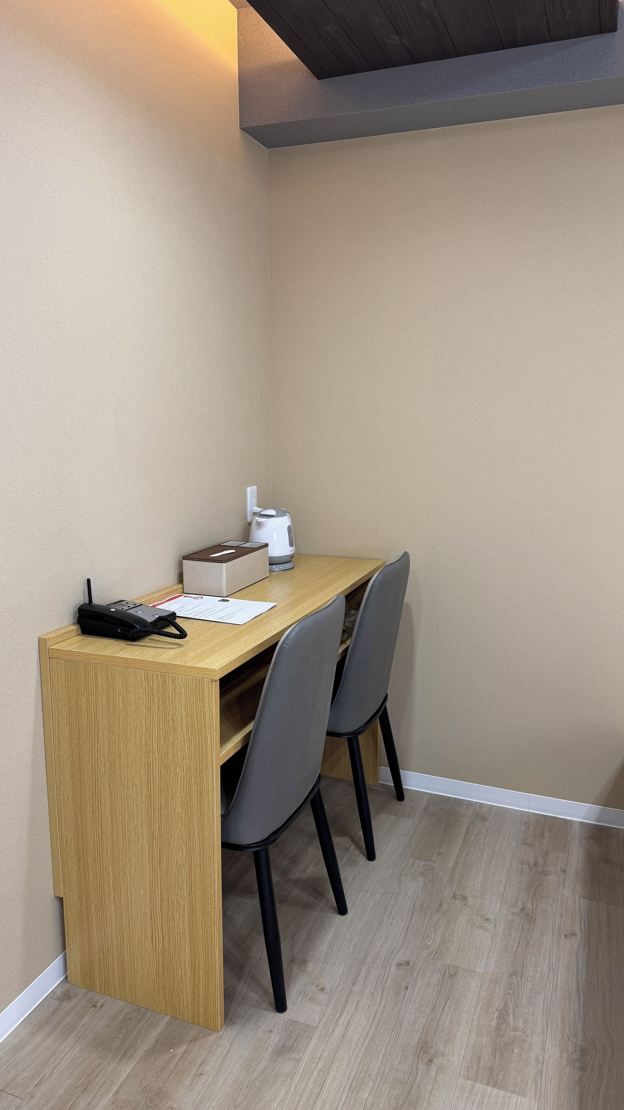 A corner room with a wooden desk, two chairs, a telephone, a kettle, and a box.