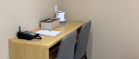A corner room with a wooden desk, two chairs, a telephone, a kettle, and a box.