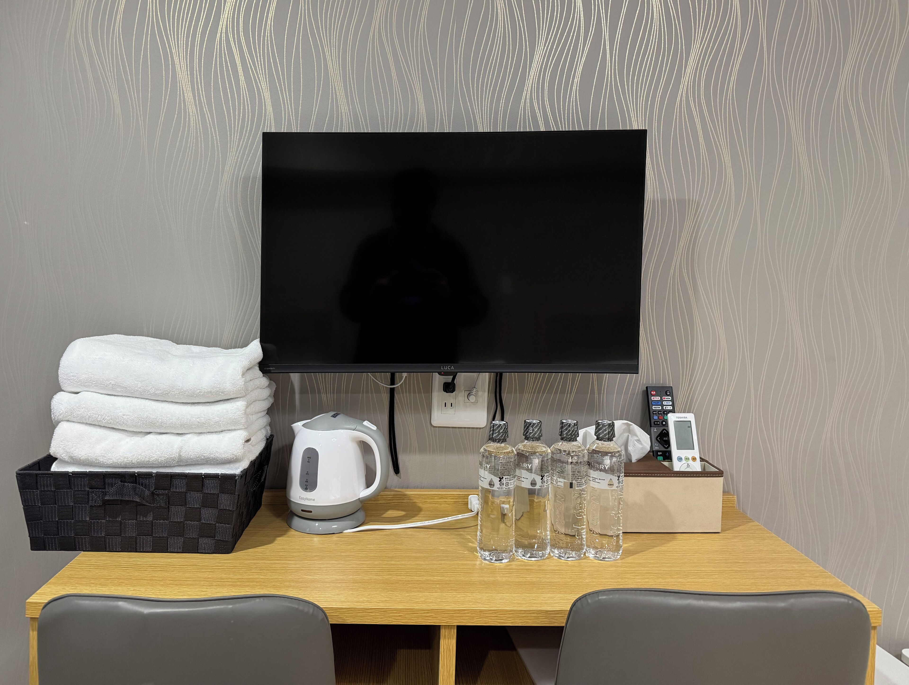 Une chambre d’hôtel équipée d’un téléviseur à écran plat, d’une bouilloire, d’une pile de serviettes et de quatre bouteilles d’eau posées sur une table en bois.
