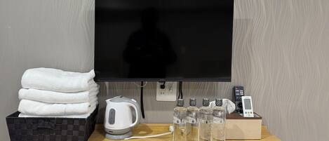 A hotel room with a flat-screen TV, a kettle, a stack of towels, and four water bottles on a wooden table.