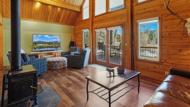 A cozy cabin interior with a fireplace, comfortable seating, a television, and large windows offering scenic views.